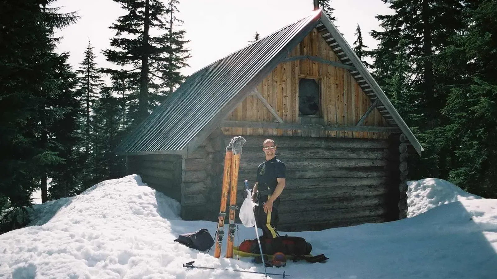 A log cabin with a steep corrugated metal roof banked in deep snow among conifers, with skis, poles and packs set out in front and a skier standing at the wall.