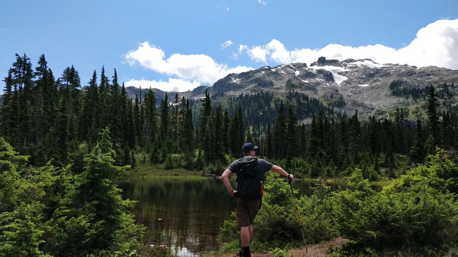 A hiker standing beside a small tarn in the upper Callaghan Valley, looking toward a snow streaked ridge above subalpine forest.