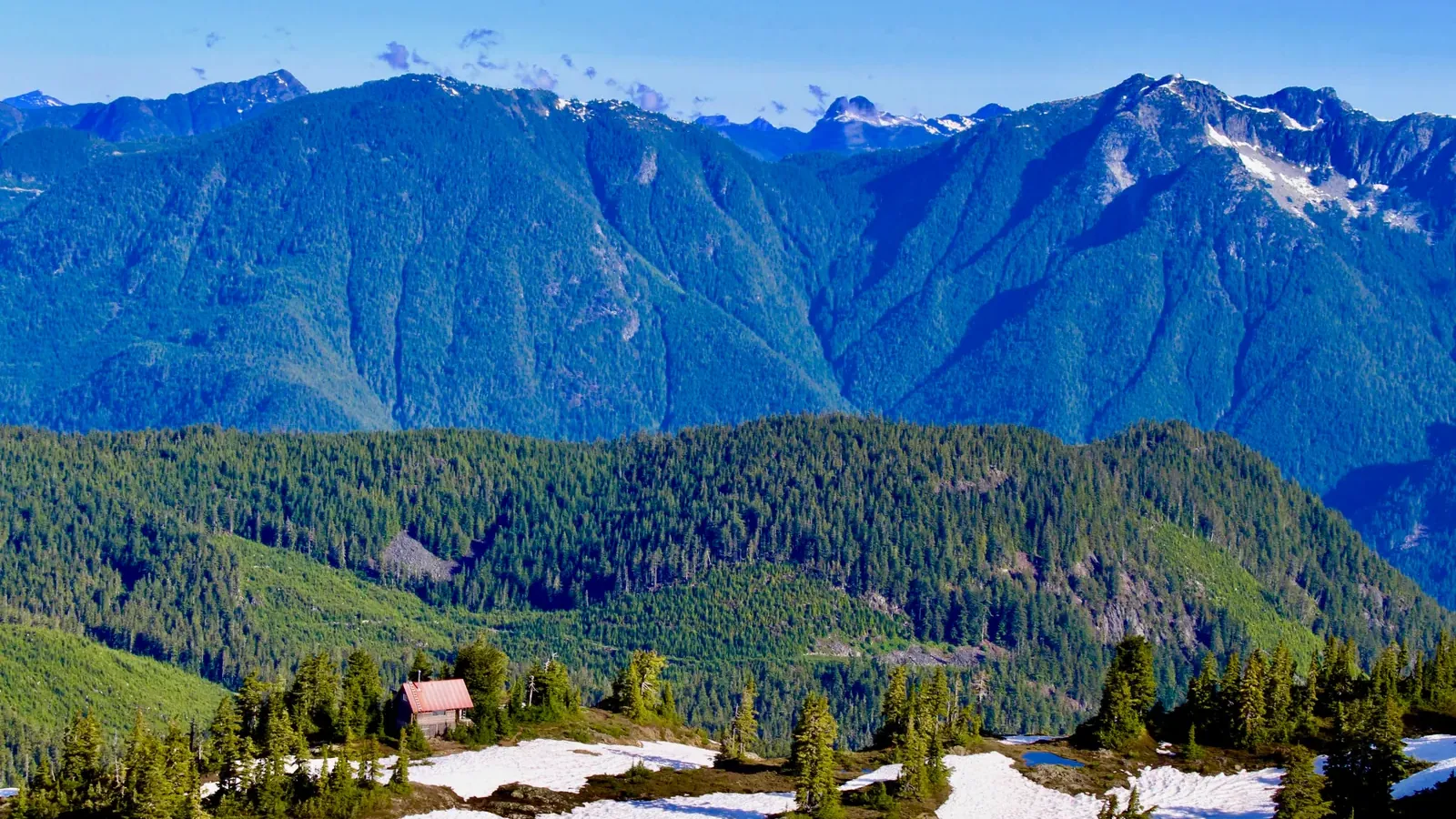 A small dark cabin on an open alpine bench with lingering snow patches and scattered conifers, forested ridges and snow-streaked peaks behind.