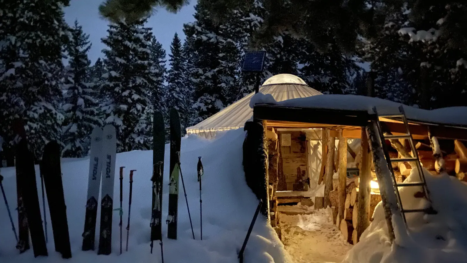 The Smith and Morehouse yurt at dusk in winter, warm light spilling from the open doorway onto packed snow, touring skis planted in a row out front and a stacked woodshed alongside